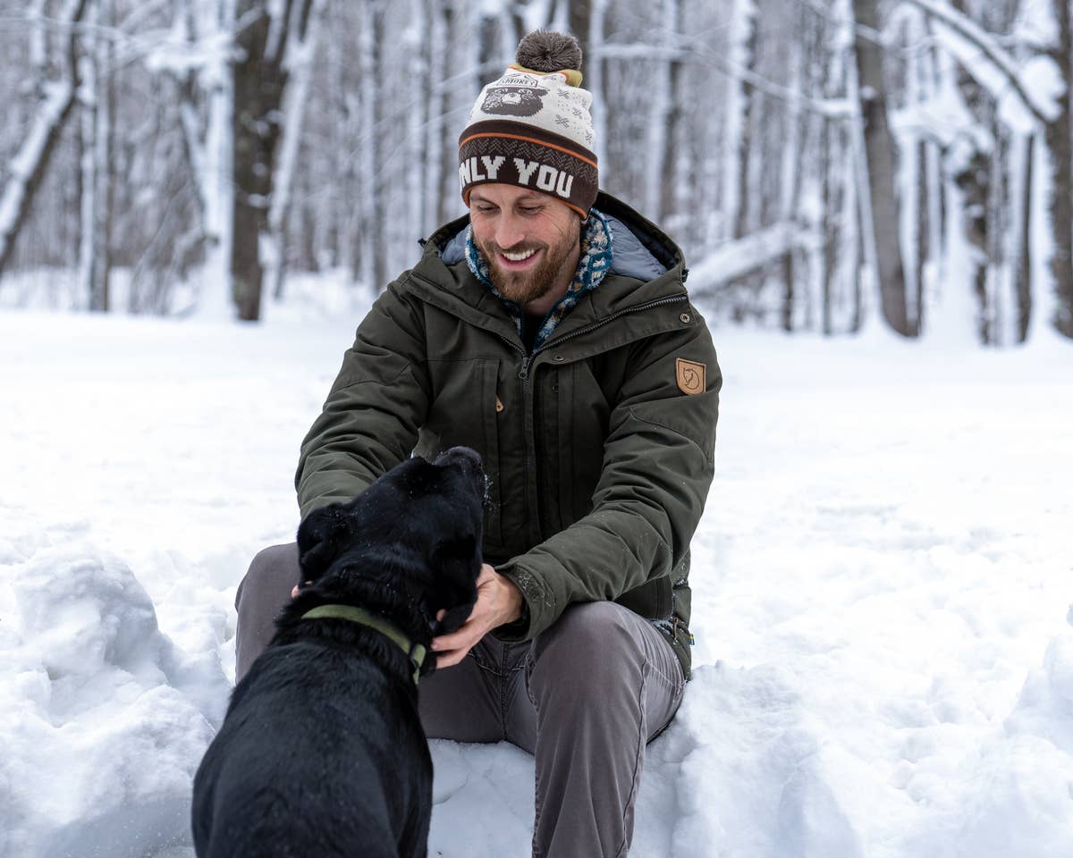 A man sits outside in the snow wearing winter clothes and a Smokey the Bear "Only You" pom beanie. He is petting a black dog and smiling.