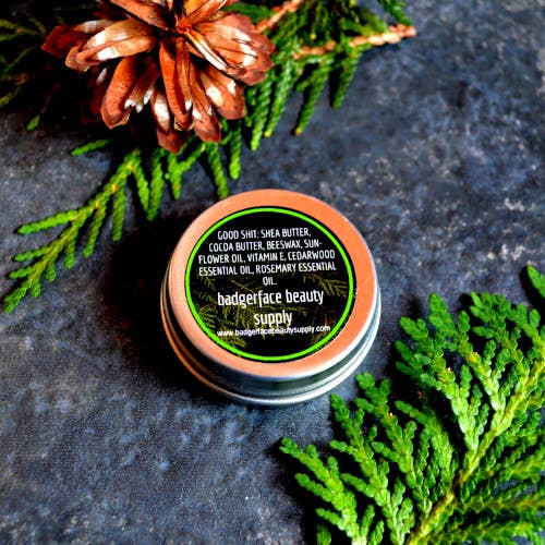 An earthy photo showing the bottom side of a tin of Badgerface Beauty Products Beard Balm. The ingredients list shows: Shea Butter, Cocoa Butter, Beeswax, Sunflower Oil, Vitamin E, Cedarwood Essential Oil, Rosemary Essential Oil.