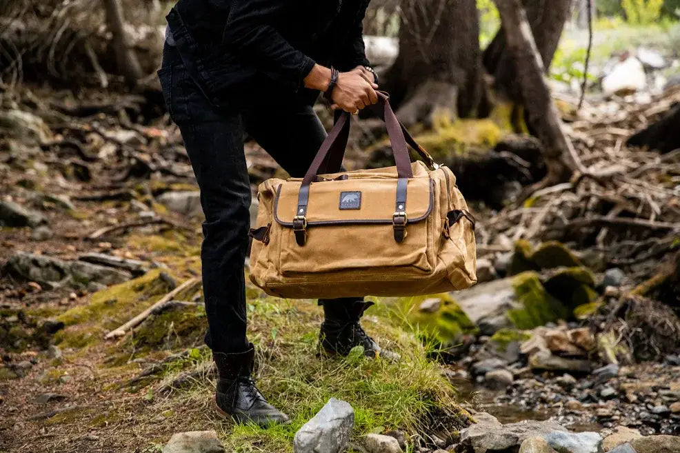 Person dressed in black holding a tan canvas and leather duffel bag by the handles, standing near a stream in the woods.