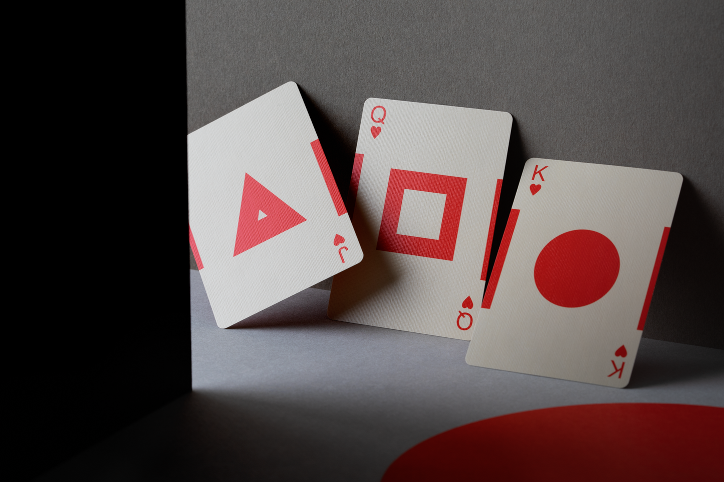 Jack, Queen, and King of Hearts cards face up showing their unique design. The Jack has a solid red triangle in the center with a small white triangle cutout of its middle. The Queen has a red border square in the middle and the King has a large solid circle in the middle. These are not traditional playing card designs.