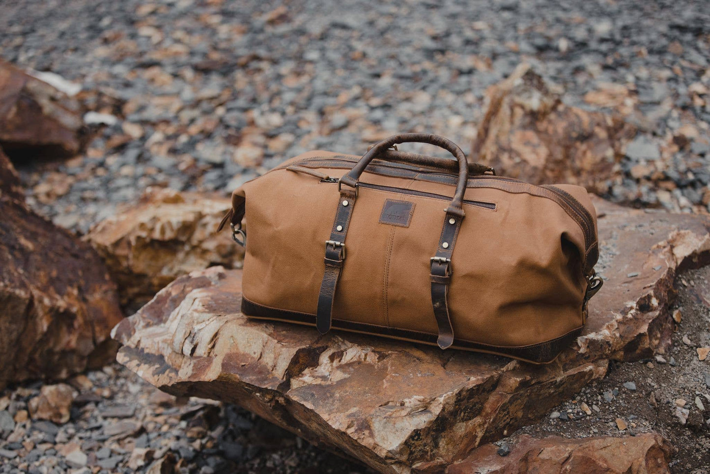 A tan canvas duffel with leather handles and straps sitting on a large rock outdoors.