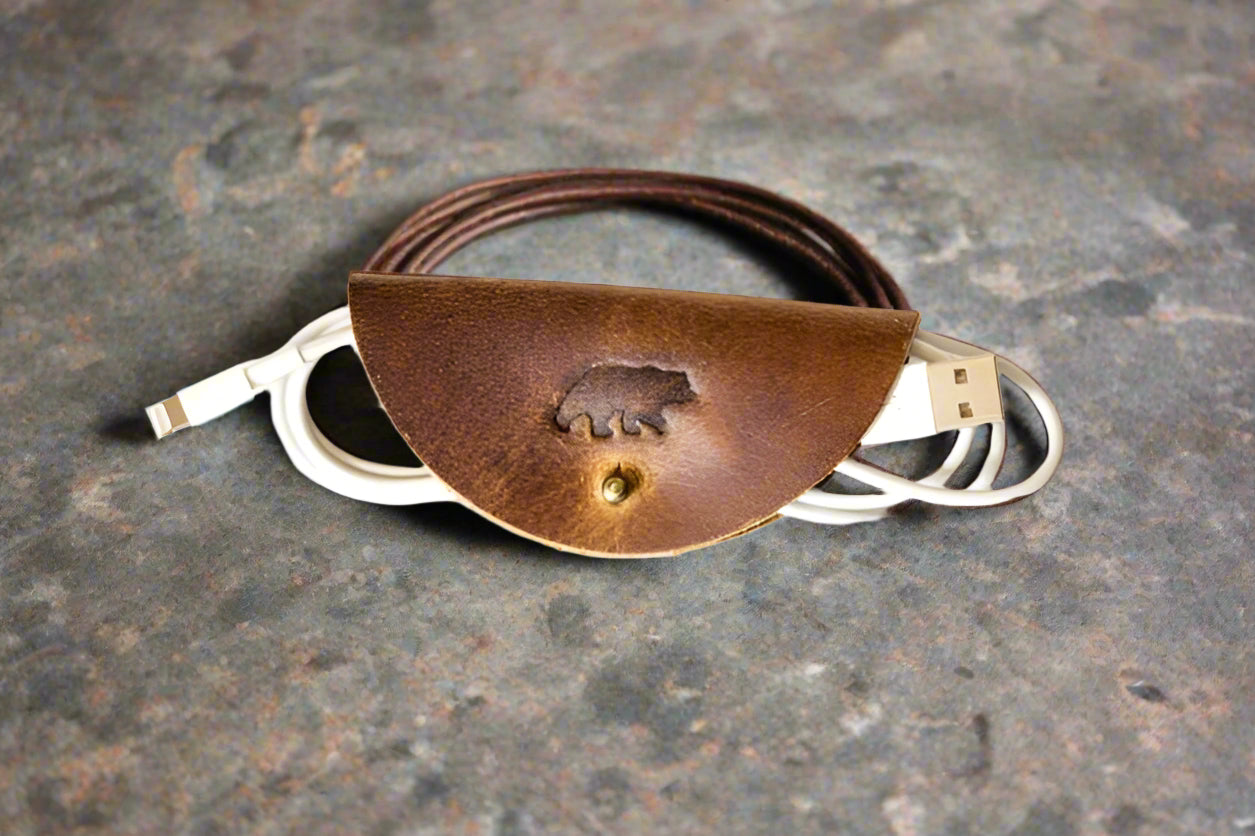 Brown leather cord wrap with an embossed bear shown on a marble surface wrapping a white and leather cord. The wrap has a snap closure.
