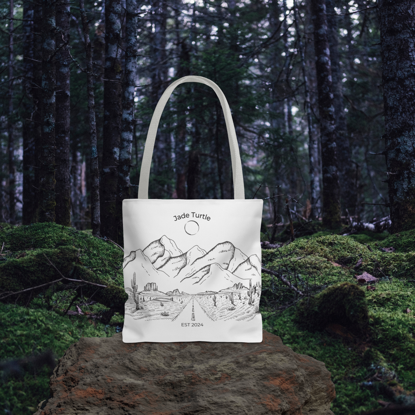 A Jade Turtle design canvas tote bag with beige handle sits on a rock in a wooded area with lots of moss on the ground covering.