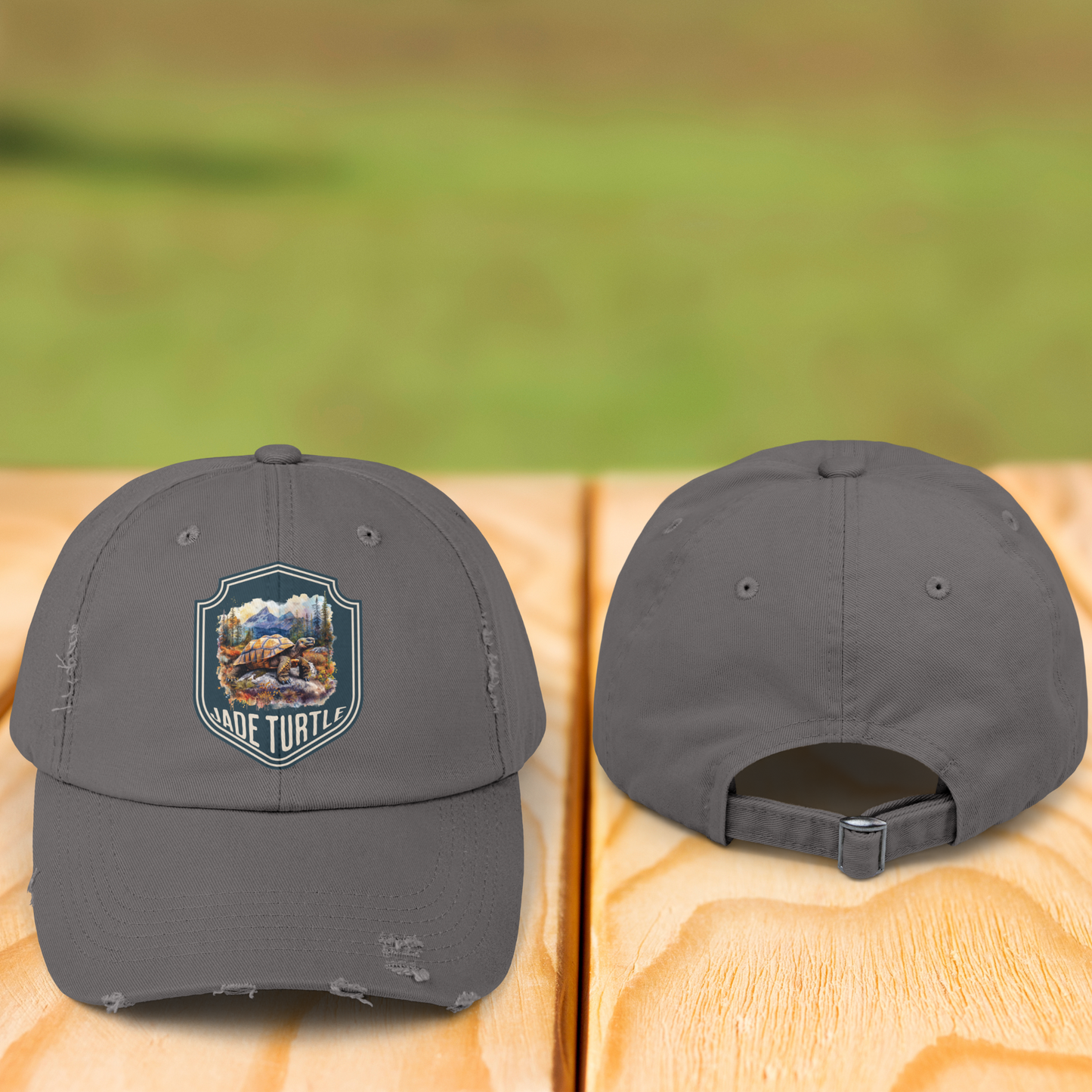 A front and back view of a gray/nickel colored distressed ball cap. The front has a Jade Turtle mountain design and they both sit on a wooden table outside with a blurred greenery background.