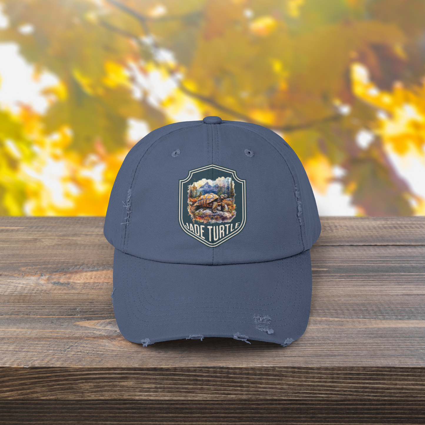 A Scotland blue distressed ball cap with a Jade Turtle mountain design sits on a wooden table outside with a blurred greenery background.