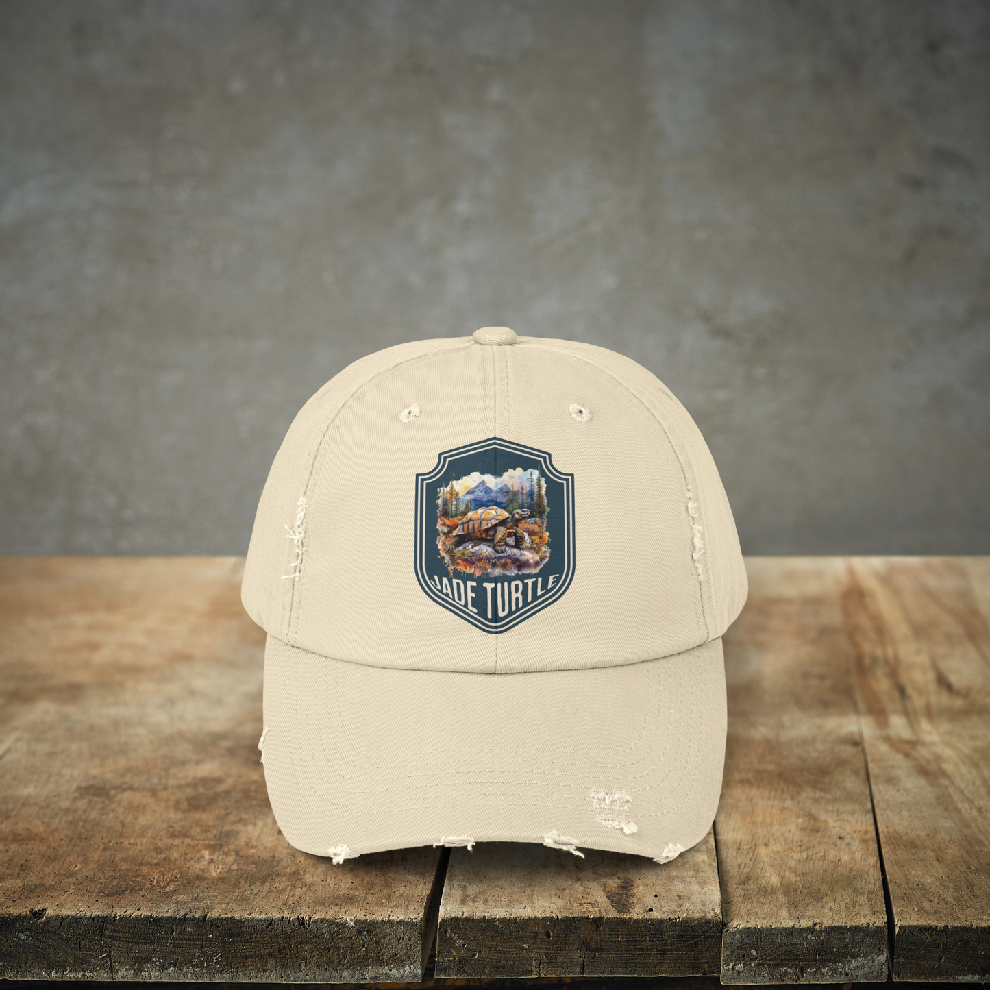 A neutral/cream/stone colored distressed ball cap with a Jade Turtle mountain design sits on a wooden table with a gray granite backdrop.