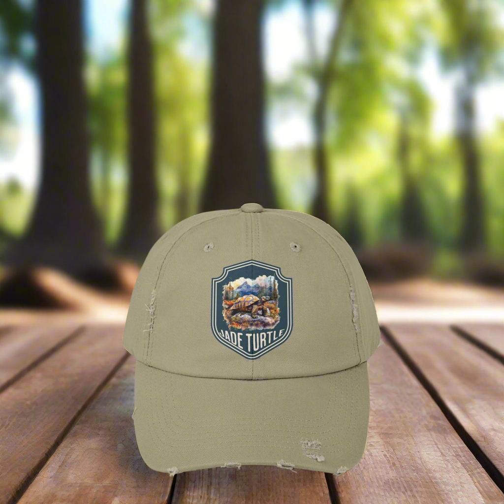 A light olive distressed ball cap with a Jade Turtle mountain design sits on a wooden table outside with a blurred greenery background.