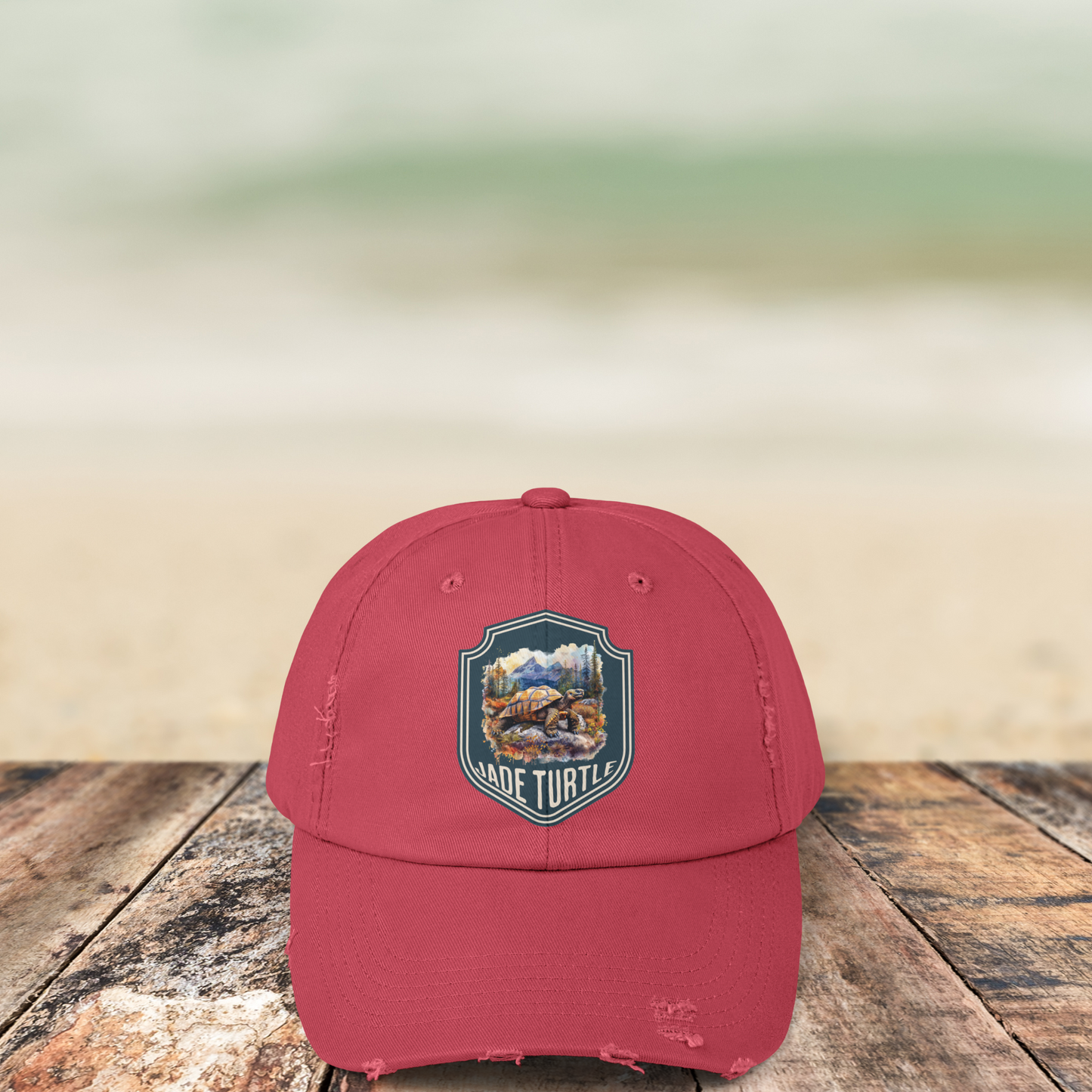 A Dashing Red distressed ball cap with a Jade Turtle mountain design sits on a wooden table outside with a blurred beachy background.