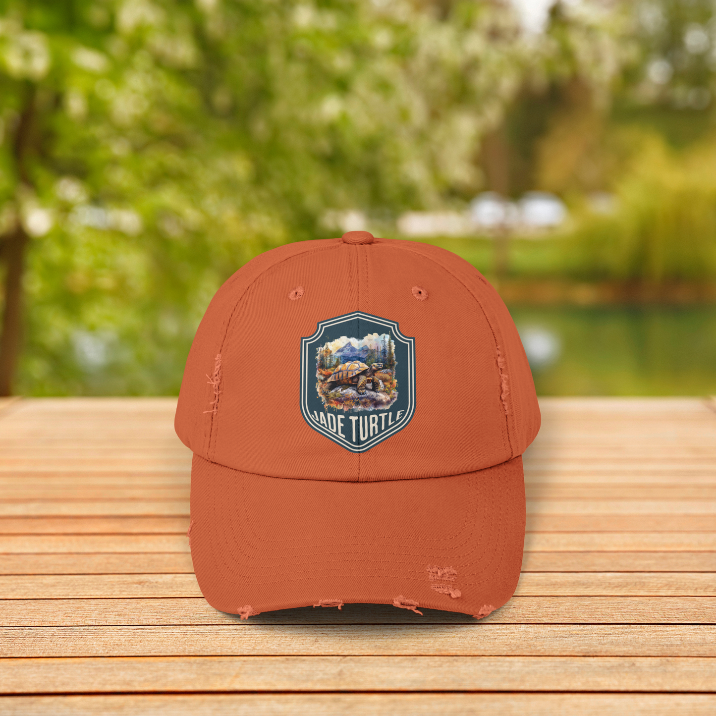A burnt orange distressed ball cap with a Jade Turtle mountain design sits on a wooden table outside with a blurred greenery background.