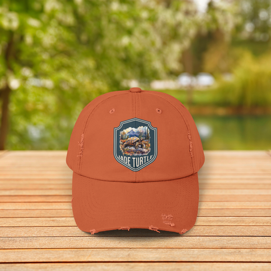 A burnt orange distressed ball cap with a Jade Turtle mountain design sits on a wooden table outside with a blurred greenery background.
