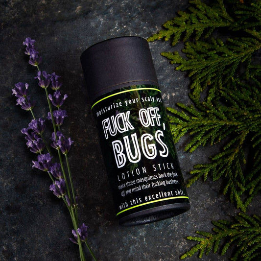 An earthy photo of a tube of Fuck Off, Bugs Lotion Stick insect repellent in plastic free container. There are cedar leaves and lavender in the photo.