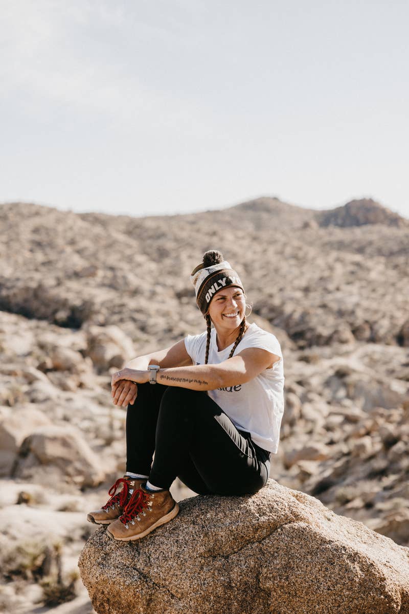 A girl with long braids and hiking clothes sits on a boulder outside. She is wearing a Smoky the Bear "Only You" pom beanie.