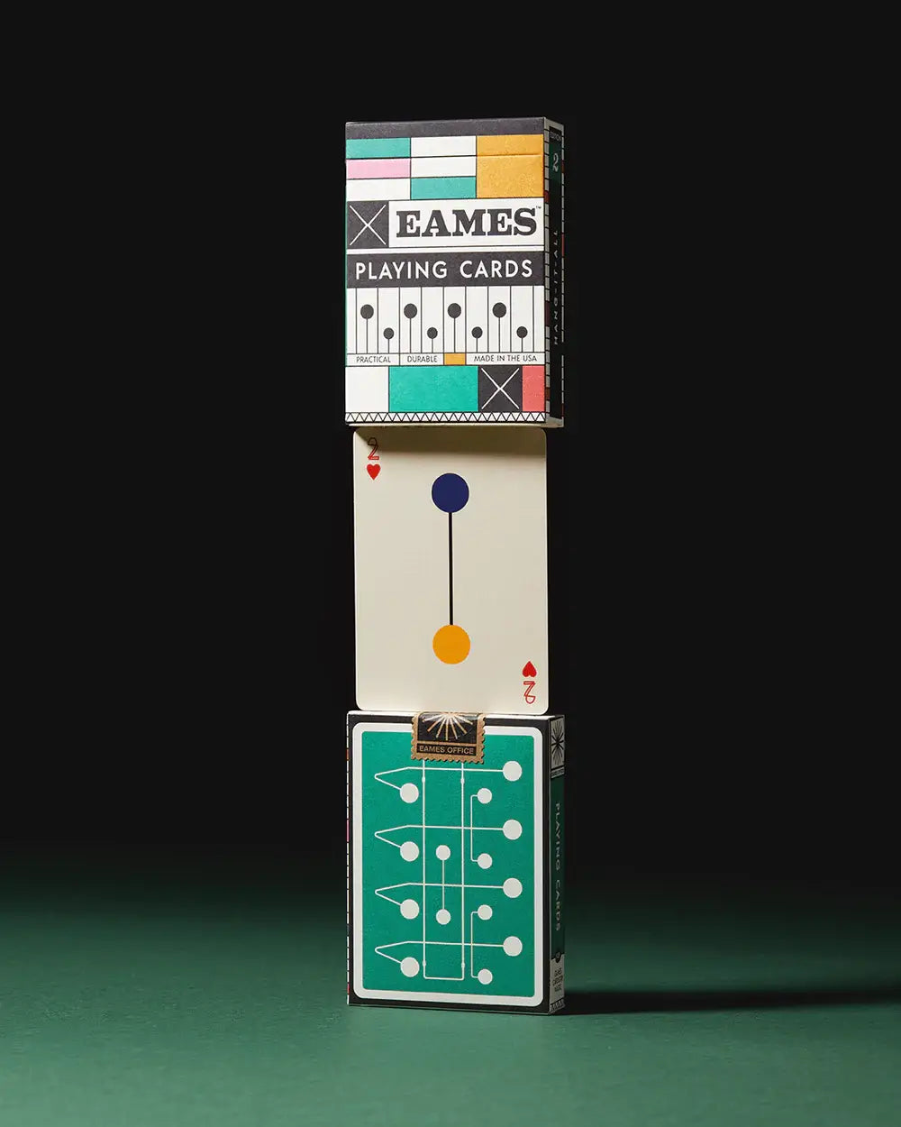 A stack of playing card deck boxes stacked end on end. One box has a green dot and line drawing. The middle is a 2 of hears card with a unique dot and line design. The top deck box shows the brand as Eames playing cards.