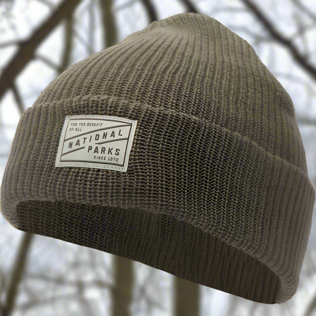 The 1872 brown National Parks beanie is shown outside with blurry winter trees in the background.