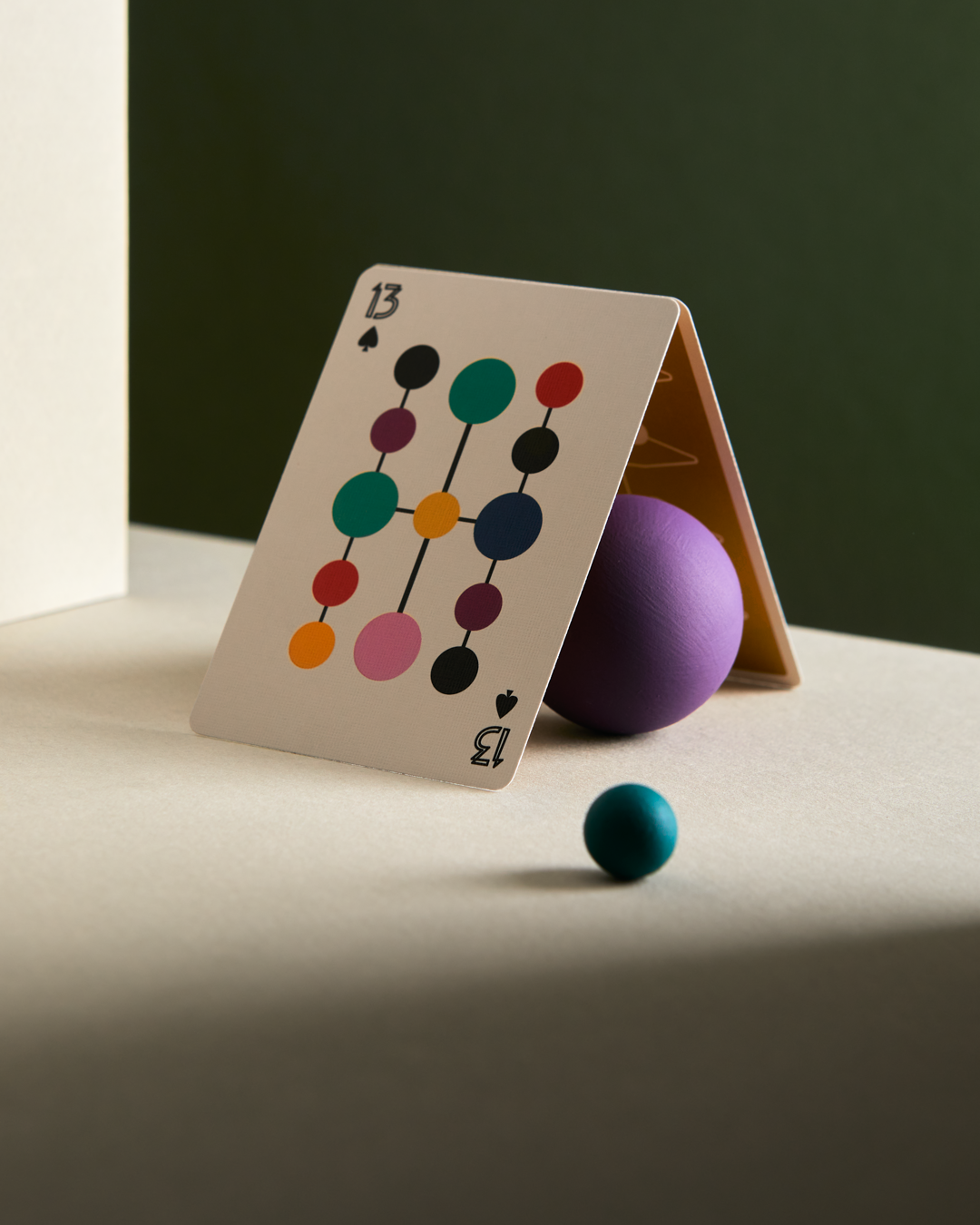 2 playing cards leaning against each other making a pyramid. The face of the showing card is a 13 of spades. A 13 instead of a king and there are thirteen colorful dots of 2 different sizes connected by straight lines.