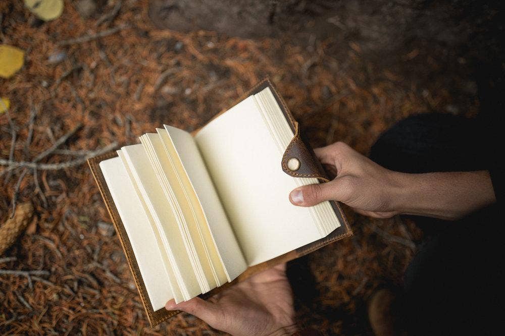 A man's hands holding an open leather journal with blank pages. The man is outside.