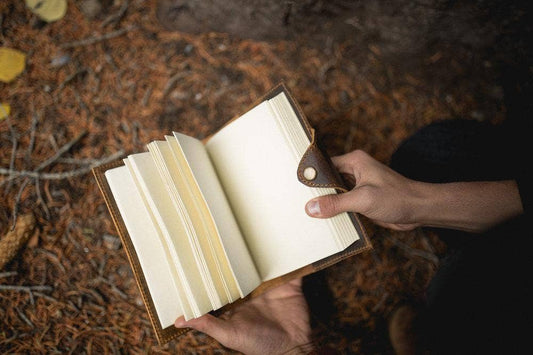 A man's hands holding an open leather journal with blank pages. The man is outside.