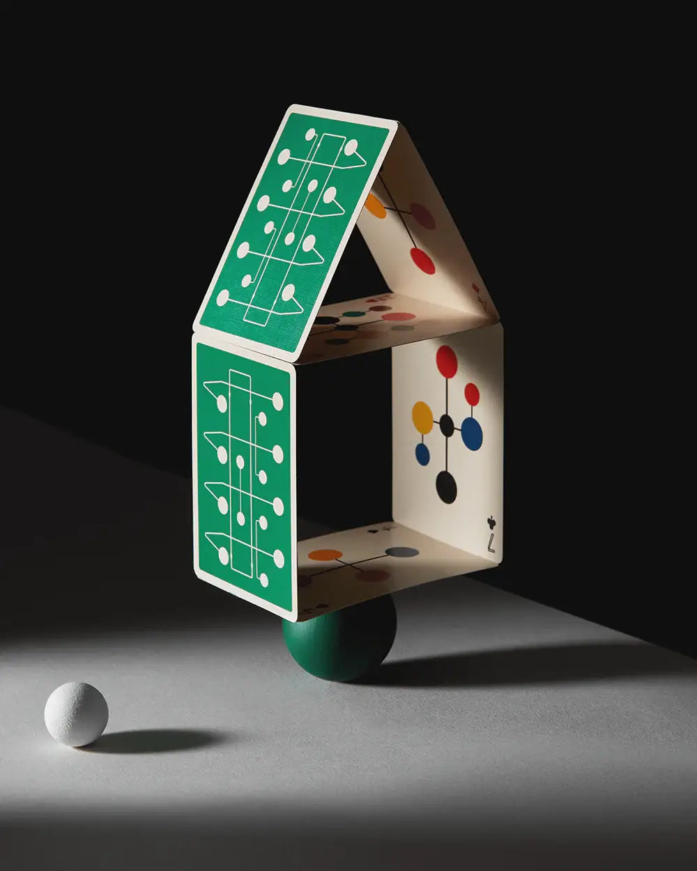 Artistic and colorful playing cards arranged in a house of cards sitting atop a green ball with dramatic lighting on the table.
