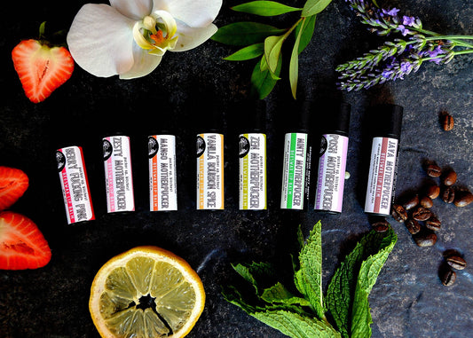 An earthy photo showing 8 flavor tubes of all natural shea butter and vitamin e lip balms. The flavors shown are: Berry Fucking Pink, Zesty Motherpucker, Mango Motherpucker, Vanilla Bourbon Spice, Zen Motherpucker, Minty Motherpucker, Posh Motherpucker and Java Motherpucker. There are also strawberries, an orange slice, mint leaves, lavender, coffee beans and a white flower shown in the photo.