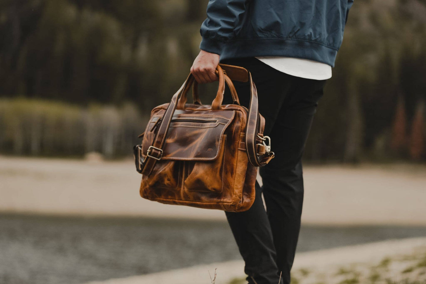 Person outside holding leather pilot bag by handles with shoulder strap showing.