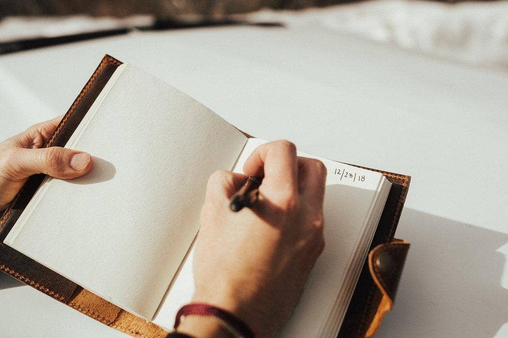 A person's hands holding a leather journal open while writing on one of the blank pages.