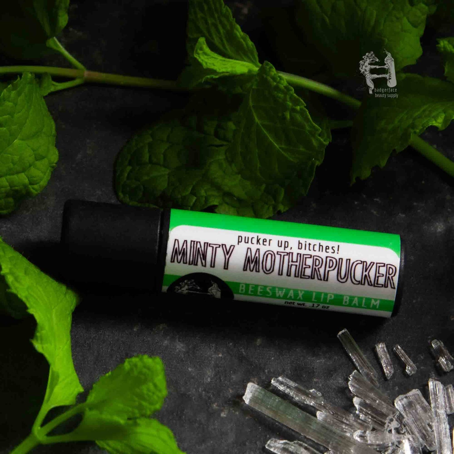 An earthy photo showing sprigs of mint with a tube of Minty Motherpucker lip balm. The label text shows, "Pucker up, bitches!"