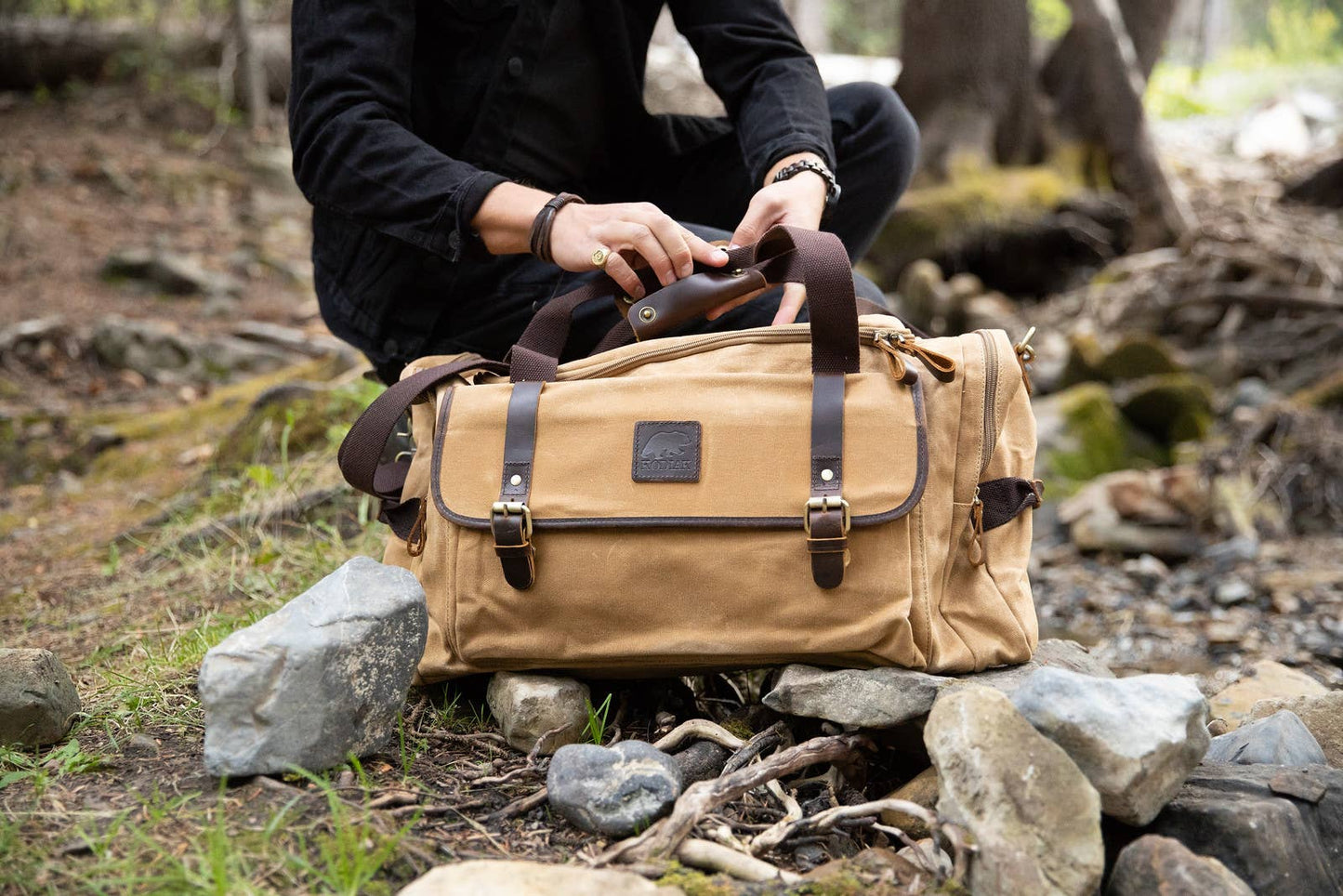 Person dressed in black closing the strap snaps on a tan canvas and leather duffel bag outdoors in the woods.
