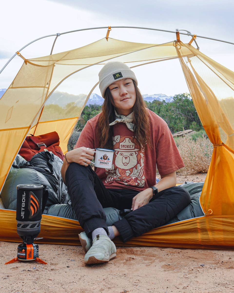 A camper with long hari and a beanie cap sits in a tent holding an enamel camping cup that has Smokey the Bear on it. The camper is also wearing a Smokey the Bear tee shirt.