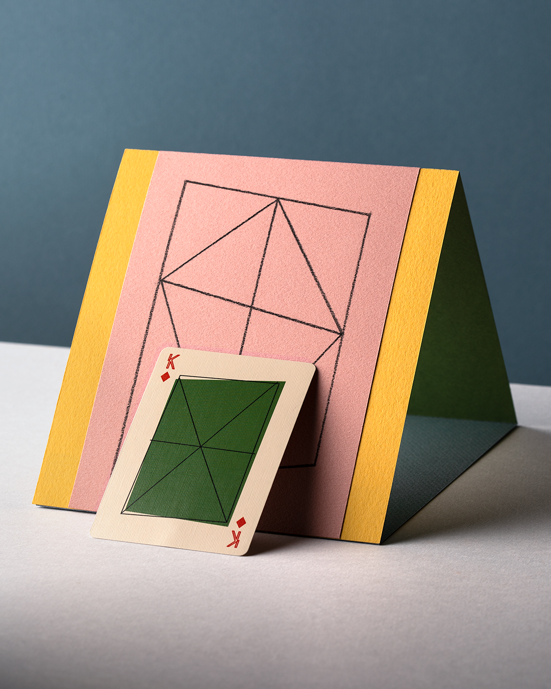 A King of Diamonds card resting upright against a paper pyramid that reflects a similar design to what is shown on the card.