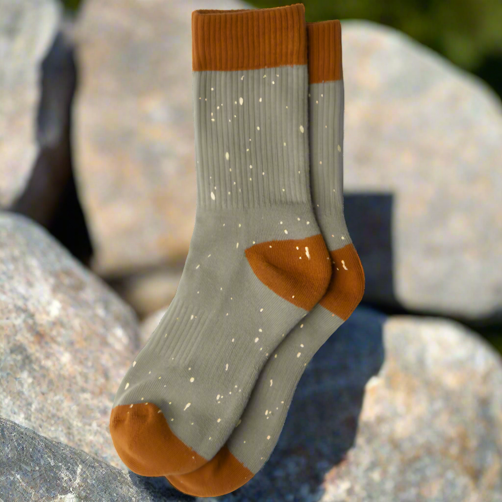 A pair of greenish gray socks with orange trim on the upper band, heel and toe area. There are "pebbles" of cream color all over. The socks are shown outside with boulder and greenery in the background.