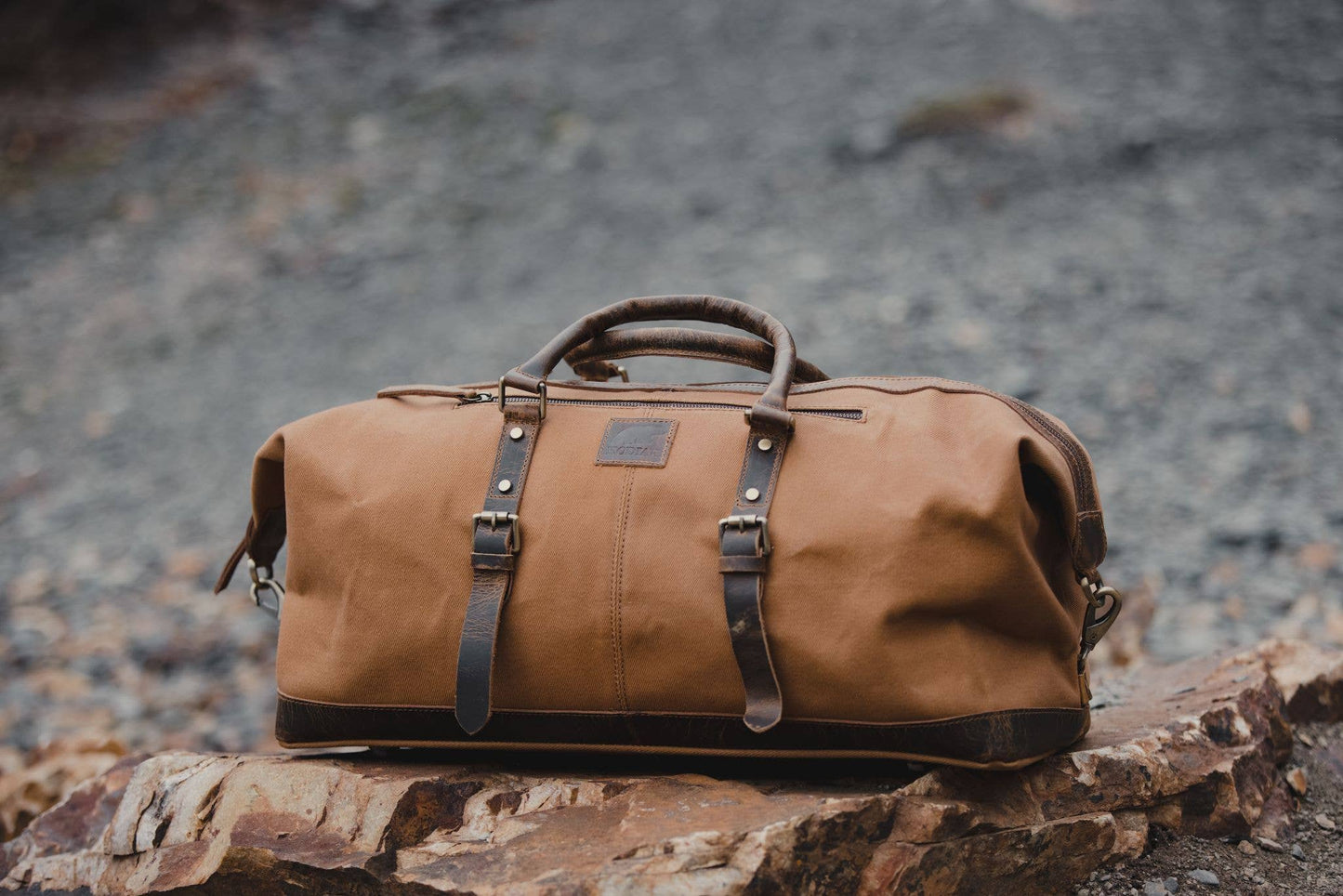 A tan canvas and leather duffel sitting on rocks outdoors.