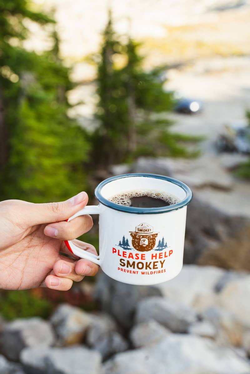 Image of a hand holding an enamel cup with coffee in it in front of a rocky overhang with trees in the background. Cup imagery is Smokey the Bear and says "Please Help Smokey precent wildfires."