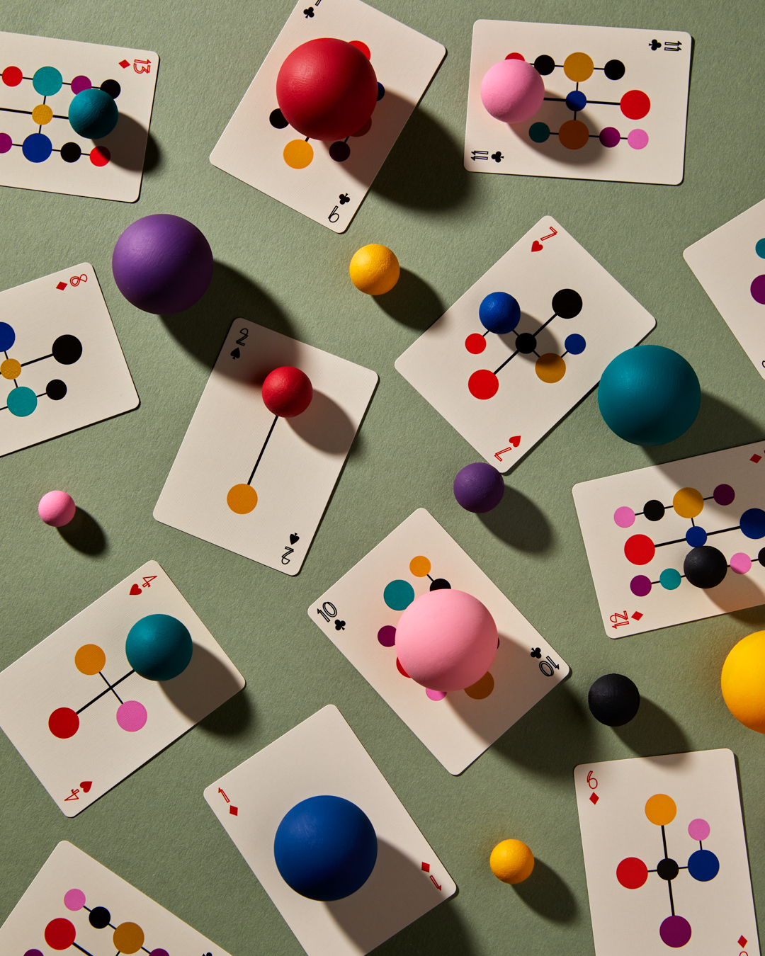 Several artistic and colorful playing cards are scattered face up on a green table. The cards show colorful dots connected by lines. A 2 card has 2 dots, a 10 card has 10 dots, etc. There are also random colorful balls of different sizes on the cards and on the table.