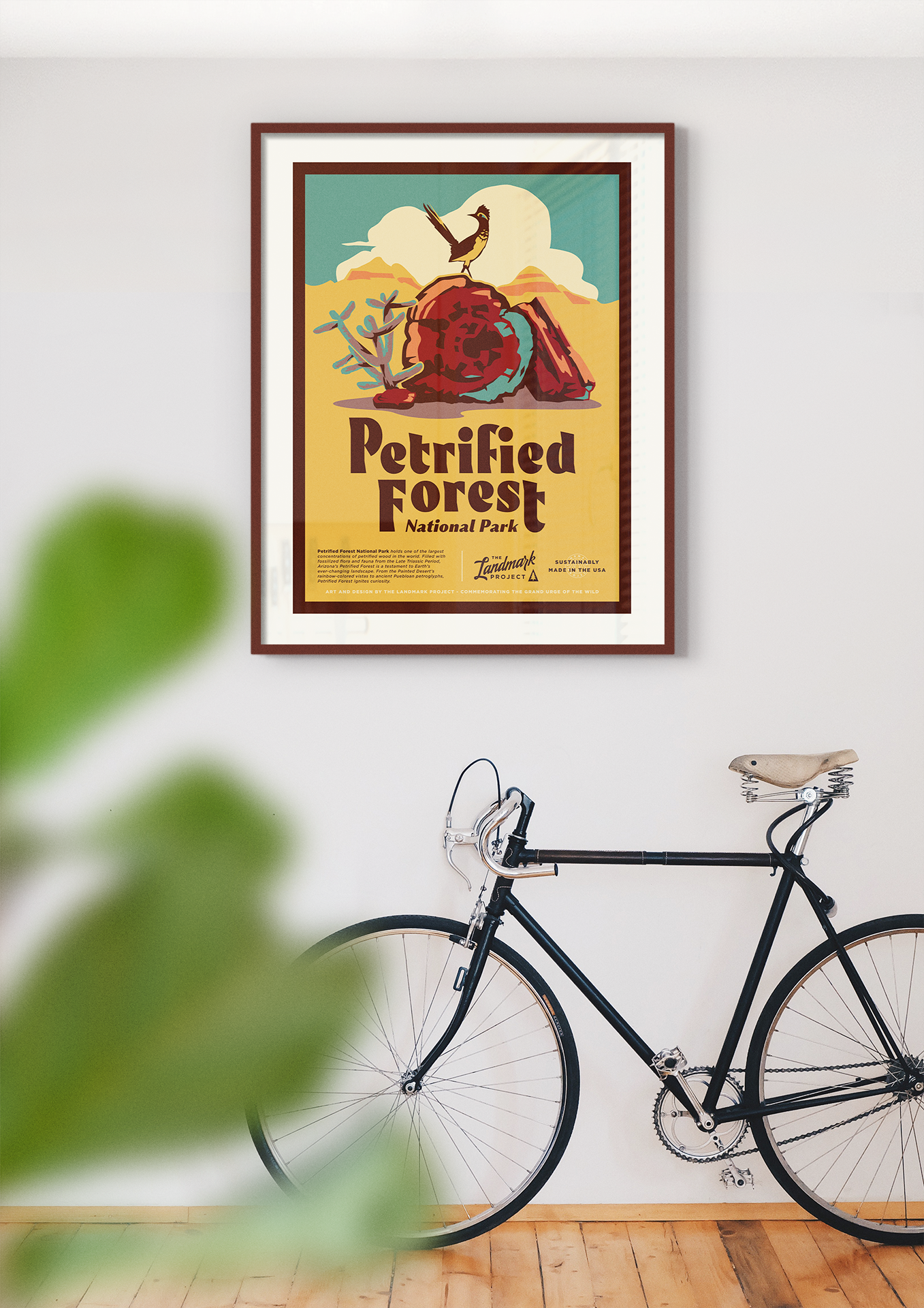 Framed Petrified Forest National Park poster is mounted on a white wall. There is a bicycle leaning on the wall below where the poster is mounted. There is greenery in the view.