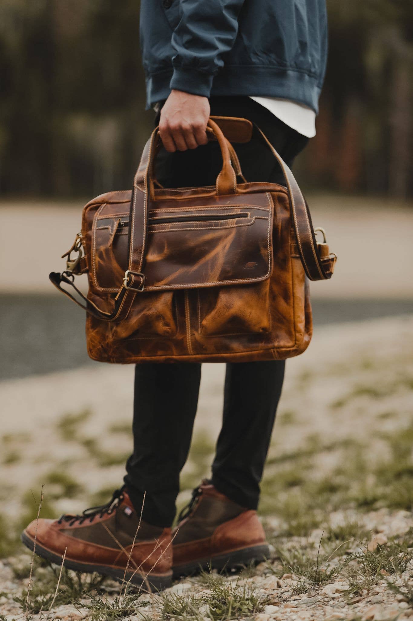 Person standing outside in the woods holding a leather pilot bag by the handles.
