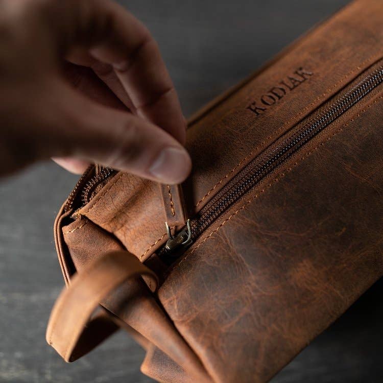 Brown leather bag with 'Kodak' branding being zipped by a hand on a dark background