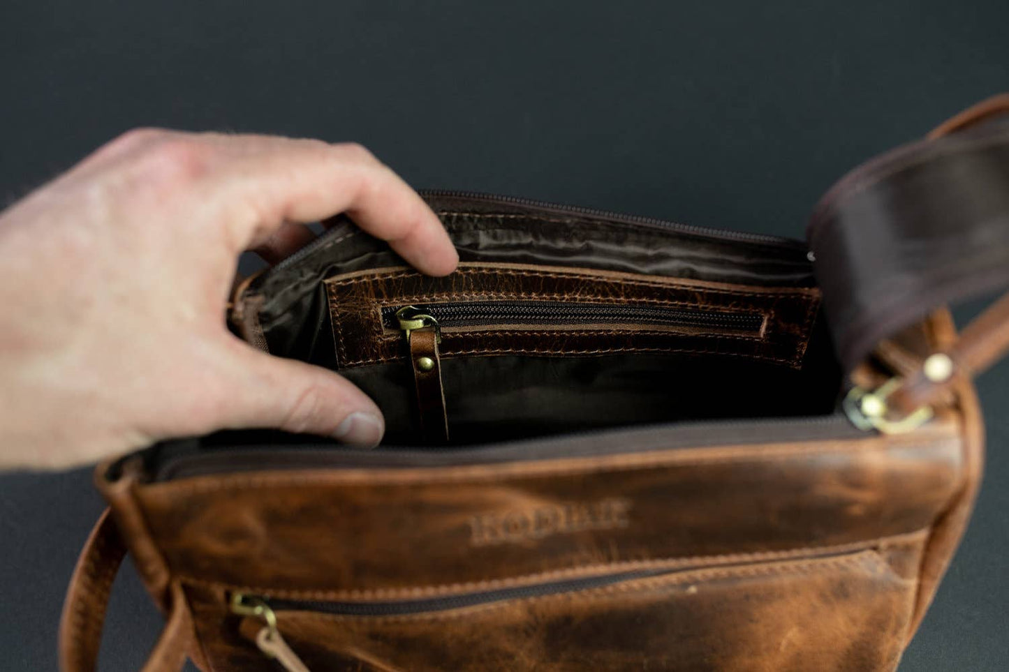 Brown leather toiletry bag with a hand opening it showing inside zipper against a dark background