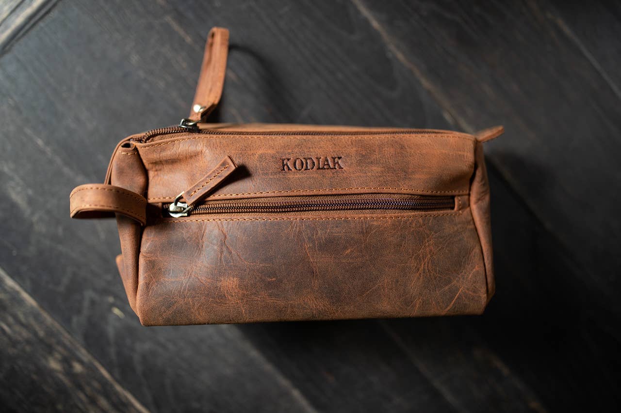 Brown leather toiletry bag with 'Kodiak' branding on a dark wooden surface