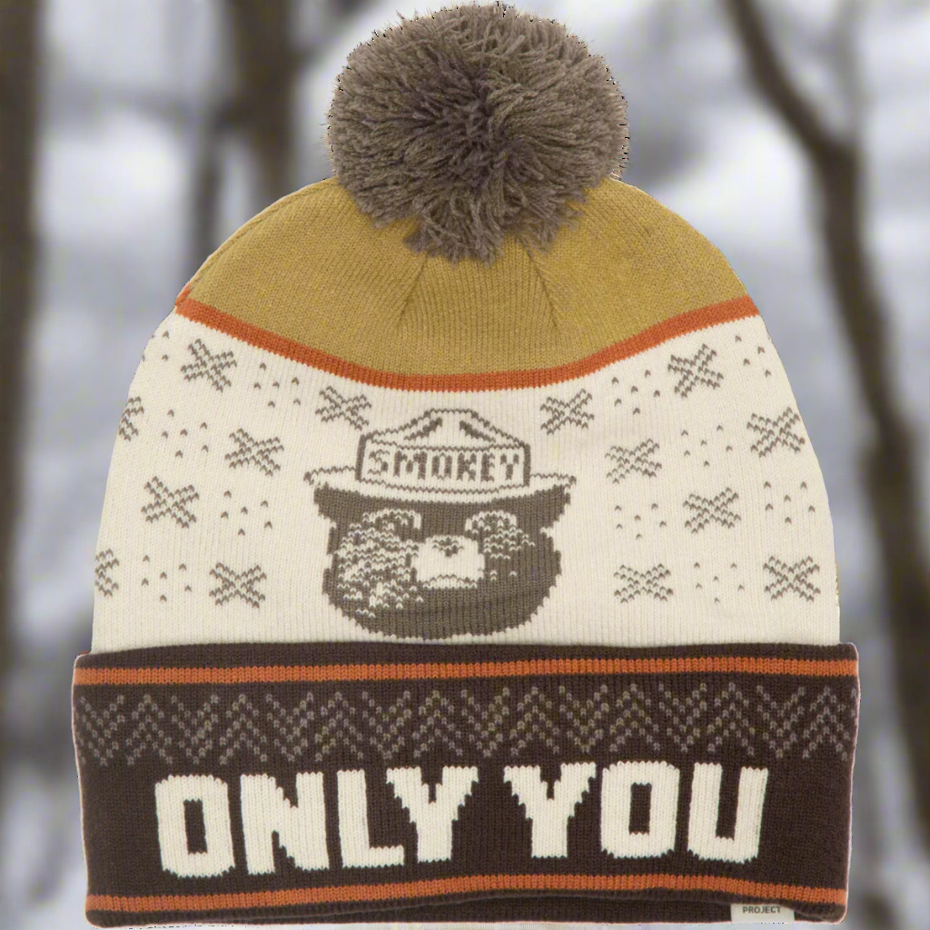 Brown, cream and gold pom beanie with Smokey the Bear image and forest design. The rolled rim says "ONLY YOU". The background is blurred winter trees.