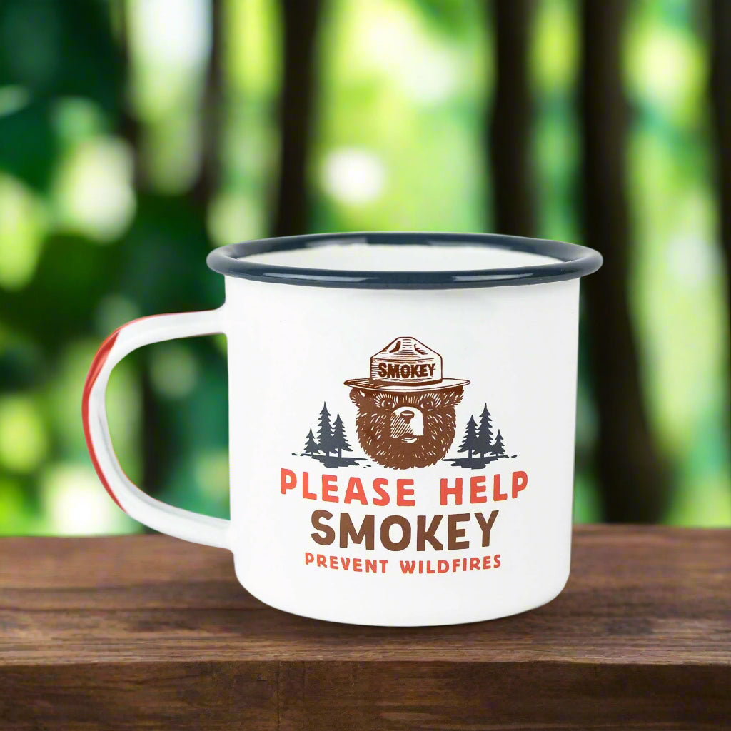 Please help Smokey camping mug on a walnut table with trees and greenery in the background. The mug has a Smokey the Bear image and says, "Please help Smokey prevent wildfires."