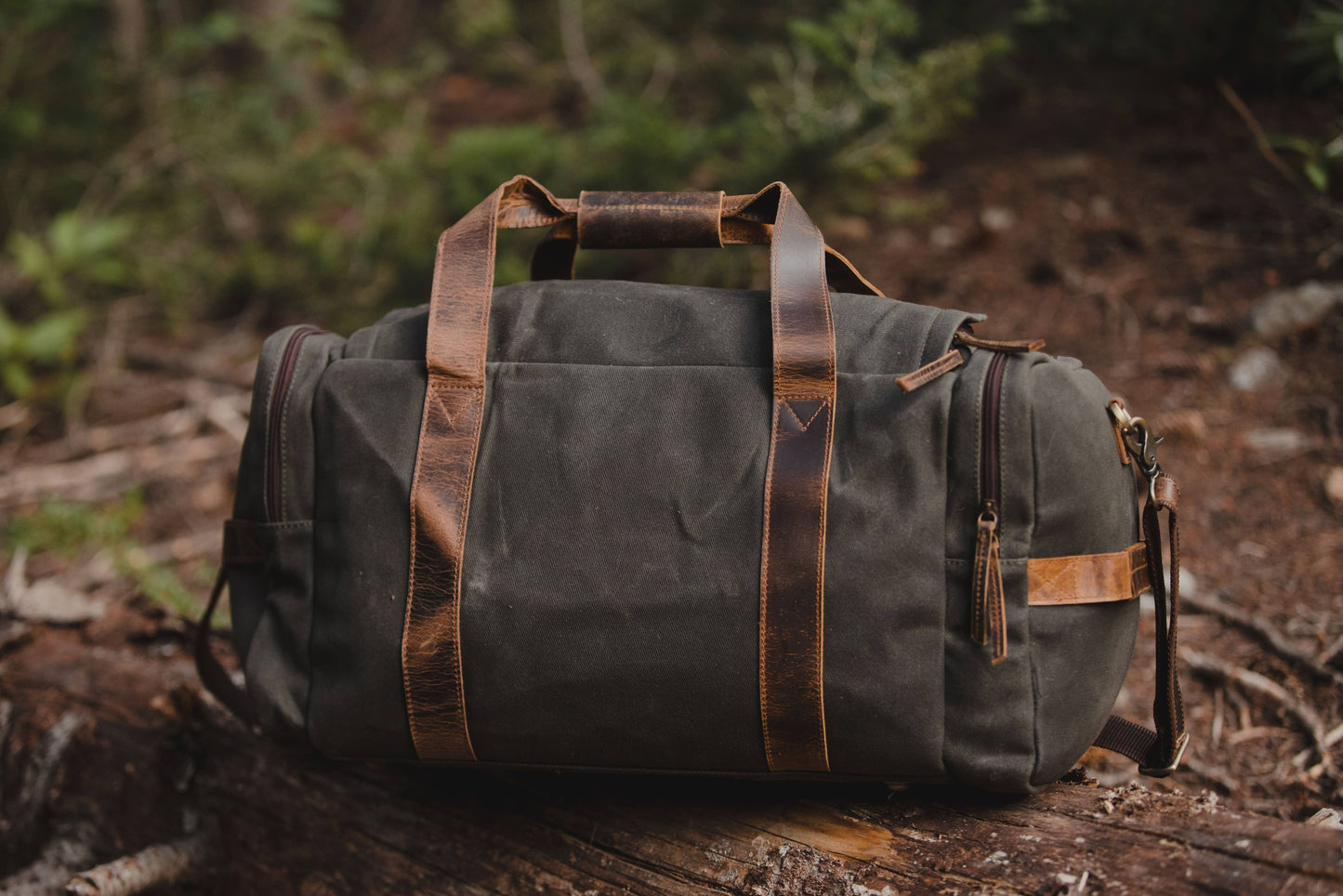 Canvas and leather duffel sitting on a log in a woodsy environment showing the leather staps.
