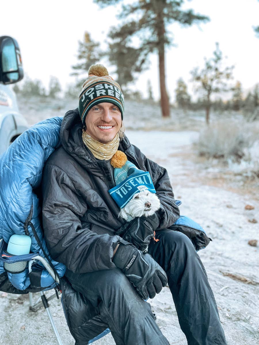 A man in winter clothing and a Joshua Tree National Park pom beanie sits in a camping chair with a white lapdog who is wearing a Yosemite beanie. They are outside in a wooded arear with a vehicle in the periphery of the photo.