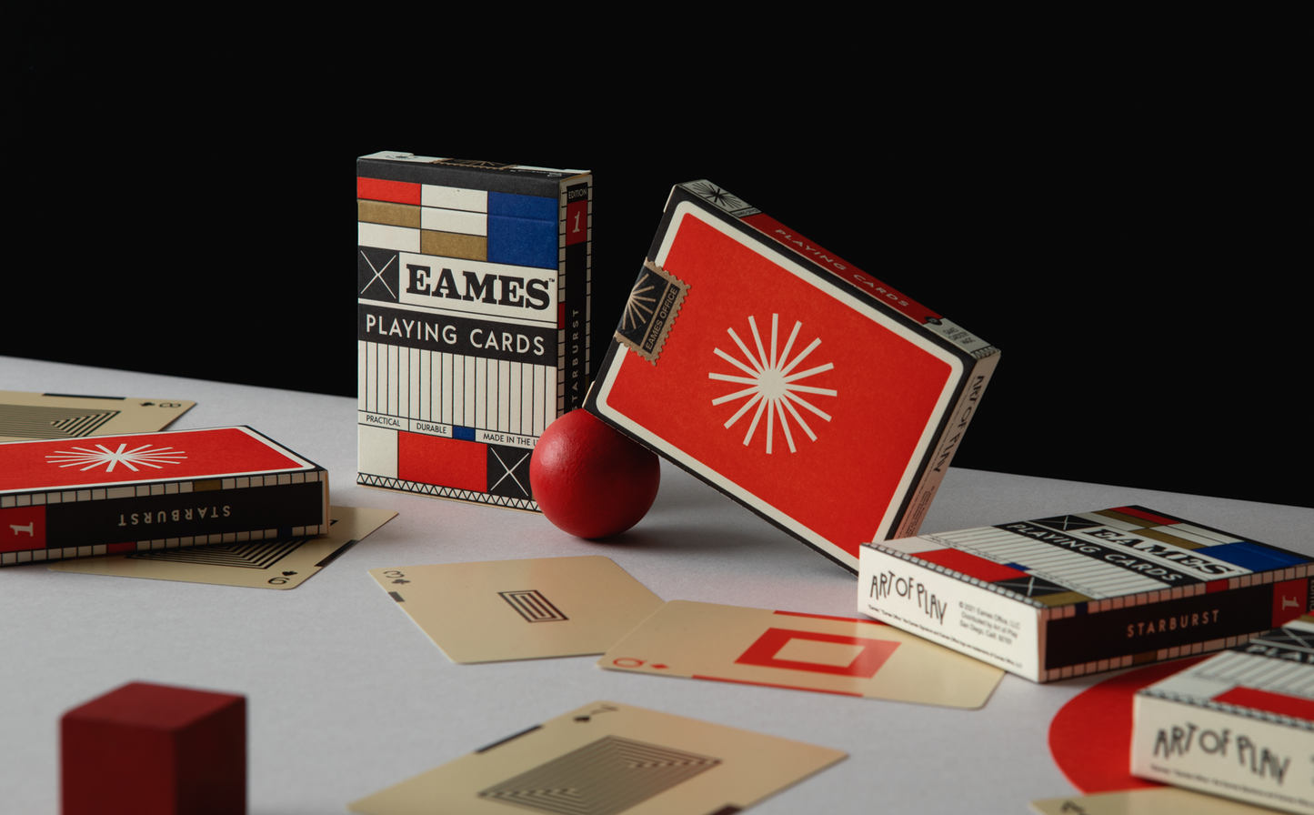 Eames playing card boxes and loose cards on a white table with a black wall behind it.