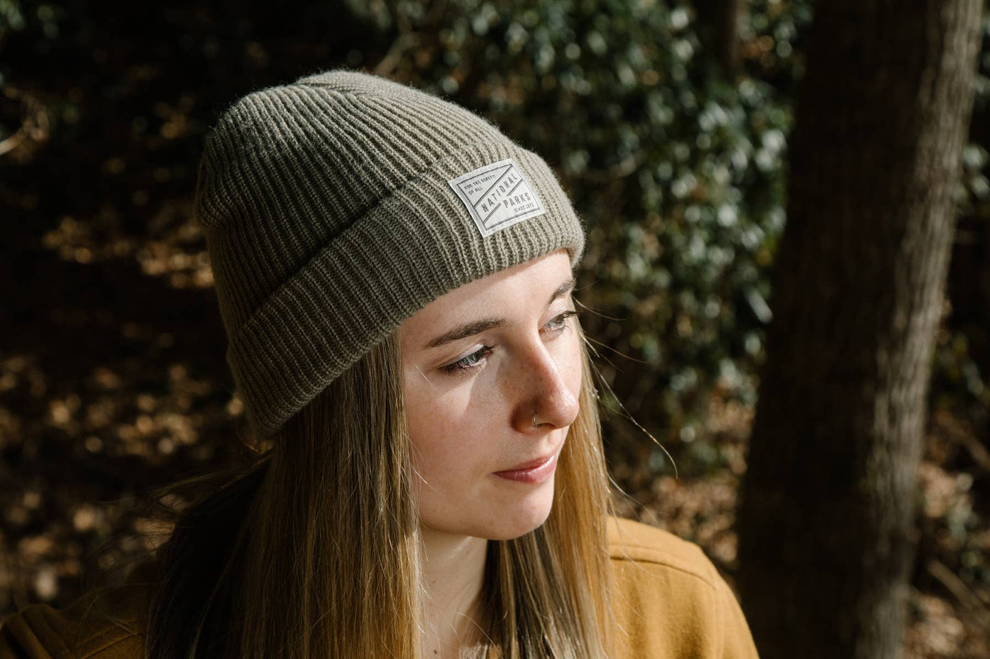 A girl with long blond hair is wearing The 1872 brown beanie outside with trees and bushes in the background indicating she is in the woods.