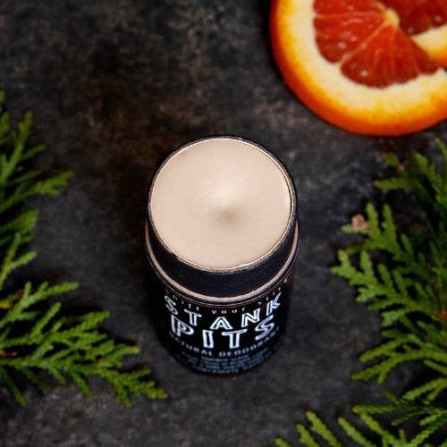 Downview photo of Stank PIts deodorant solid showing the texture with the lid off. There is a dark background with cedar and an orange slice in the photo.