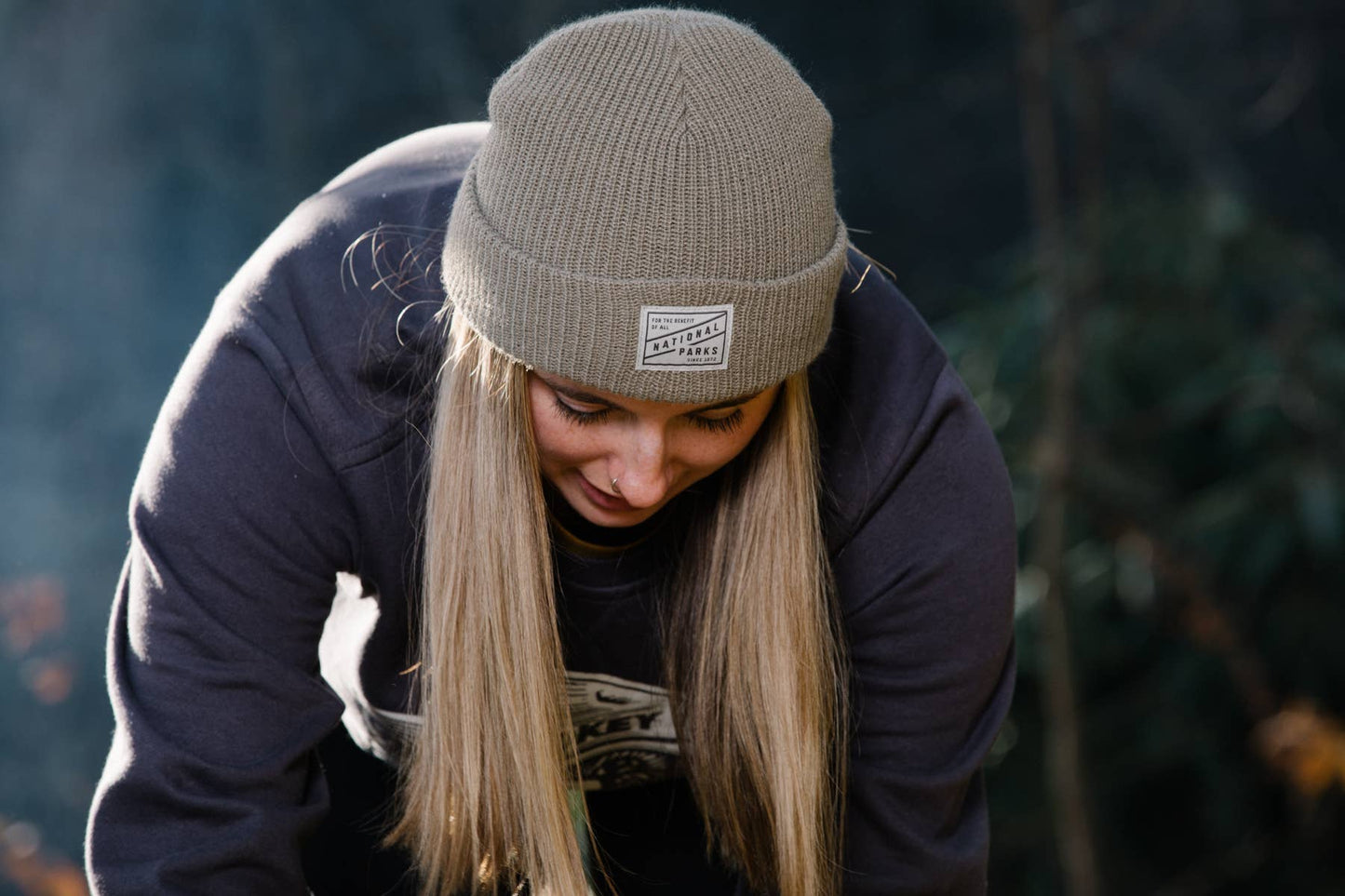 A girl with long blonde hair is outside in the woods wearing The 1872 brown beanie. She has on a thick sweatshirt and is bent over attending to something out of the picture frame.