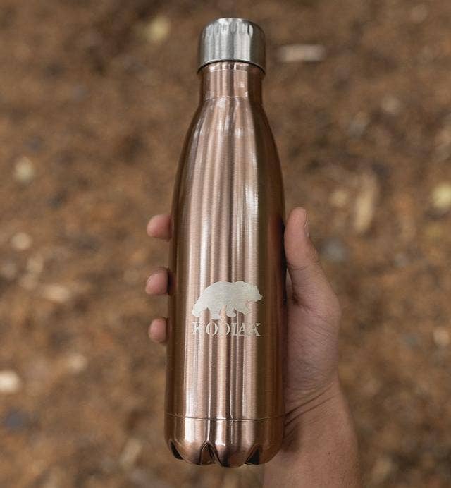A hand is holding a Stainless Steel water bottle in copper finish. The bottle has a bear and the brand name "Kodiak" etched on the side. The background appears to be outside over a forest floor.