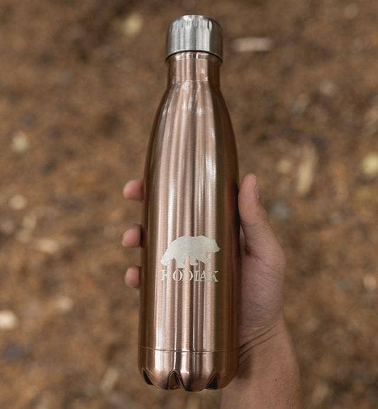 A hand is holding a Stainless Steel water bottle in copper finish. The bottle has a bear and the brand name "Kodiak" etched on the side. The background appears to be outside over a forest floor.