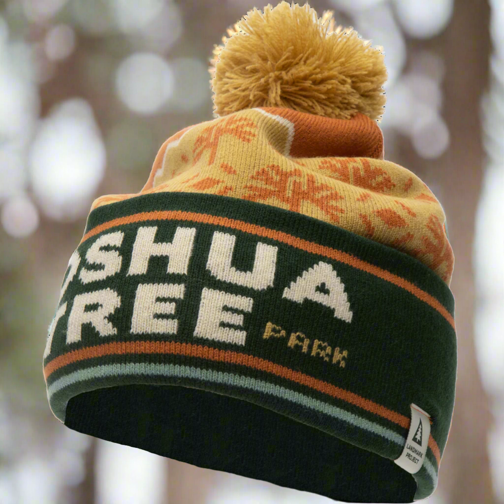 Side view of an orange and green Joshua Tree Nat'l Park pom beanie outdoors with blurred trees in the background.