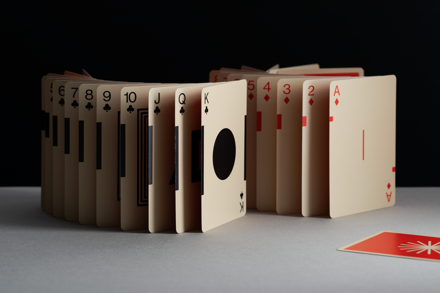 Suits of clubs and diamonds cards artfully arranged standing up like dominoes on a white table in front of a black wall.